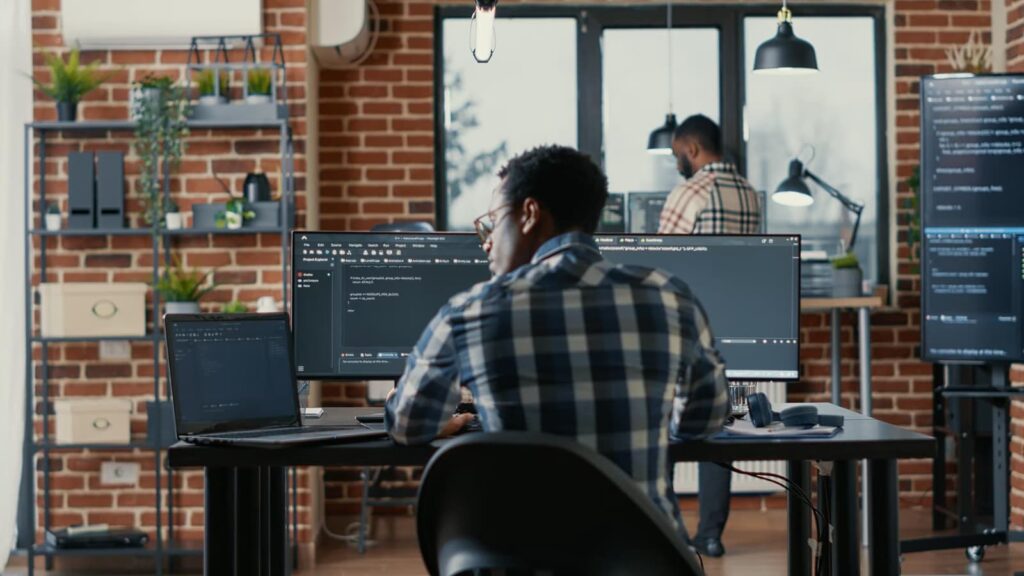 Programmer writing code on laptop, sitting at table with computer screens in software agency